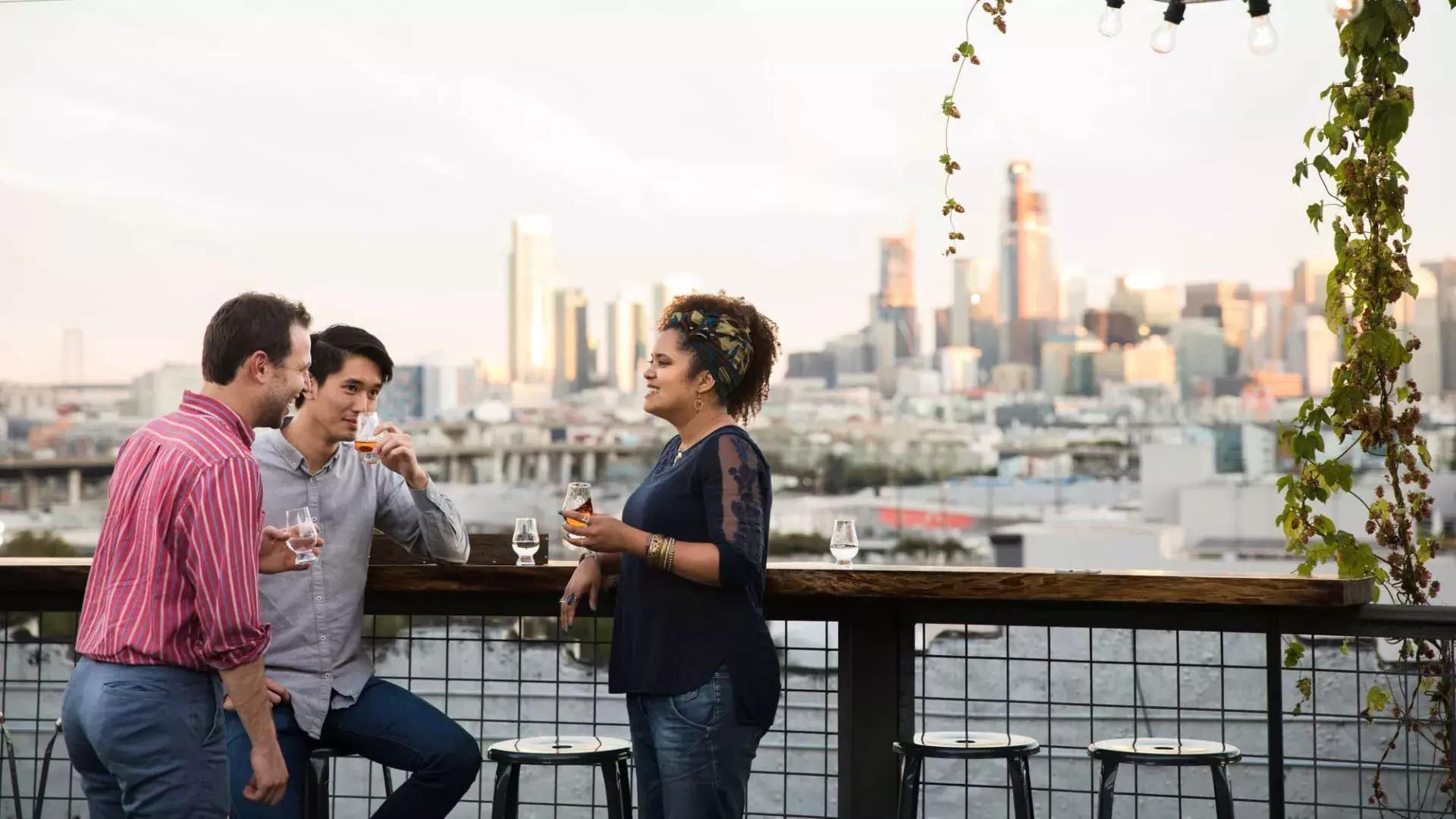 三个人围坐在贝博体彩app“锚蒸馏”(Anchor Distilling)屋顶甲板上的一张户外桌子旁, 加州.